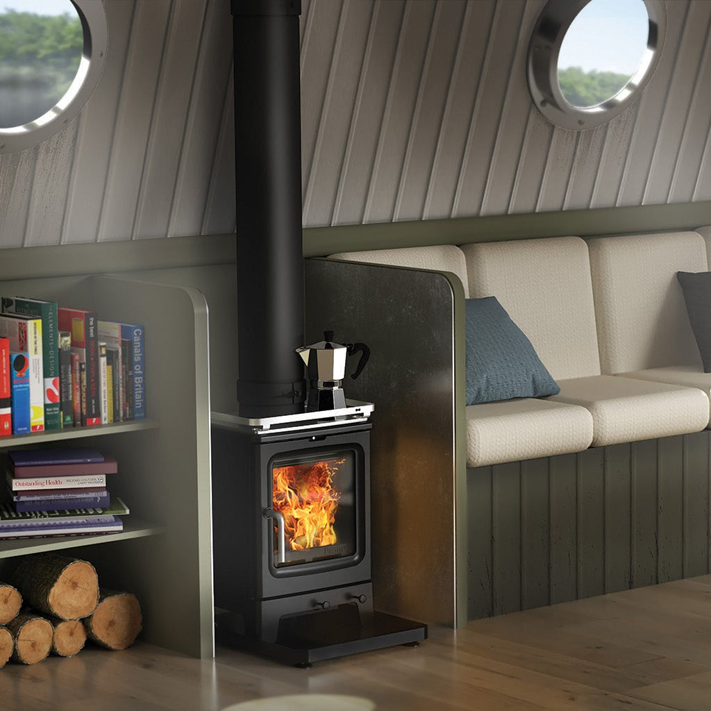 A cozy interior scene with a built-in cream sofa, gray cushions, a black wood-burning stove with visible flames, round porthole windows, shelving with books and logs, and a wooden coffee table.