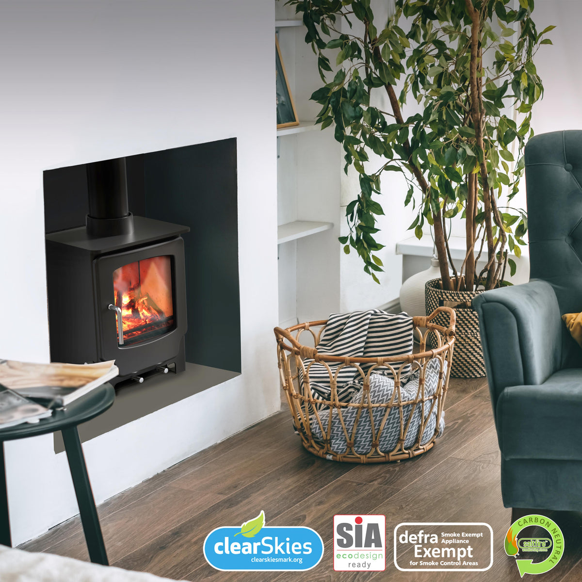 A modern living room scene featuring a black wood-burning stove installed in a white wall niche, alongside a deep green tufted armchair with a mustard yellow pillow, a potted plant, and a wicker basket. The 'SALTFIRE-SCOUT' brand logo is visible in the top right, and certification badges are at the bottom.