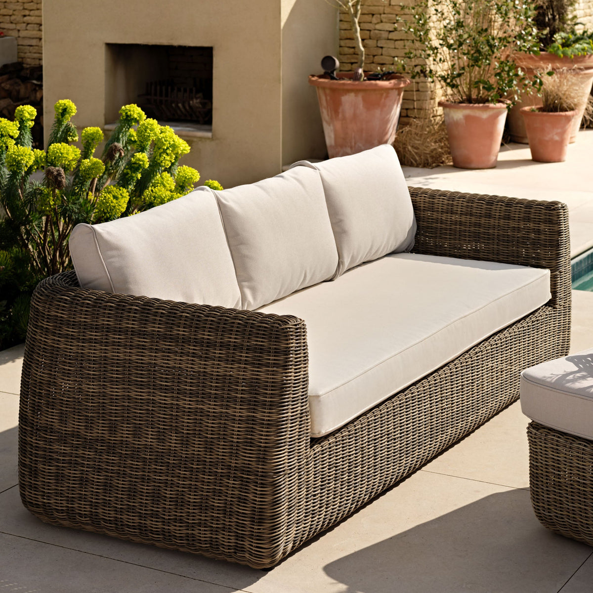 Wicker outdoor sofa with white cushions on a patio with plants and a fireplace.