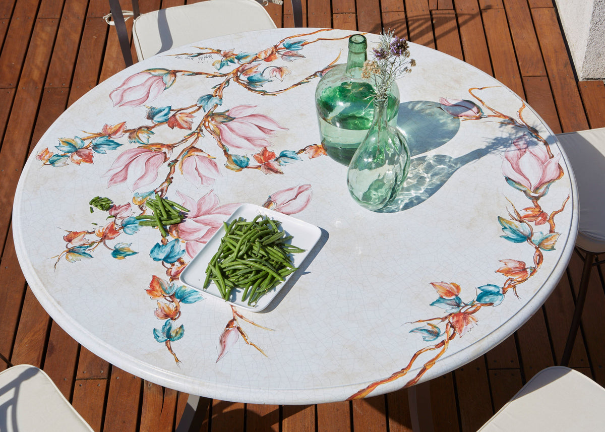 A round outdoor dining table with a white marble-look surface decorated with a colorful hand-painted floral branch pattern. The table is styled with two green glass bottles and a small dish of green beans, set on a wooden deck with white chairs.