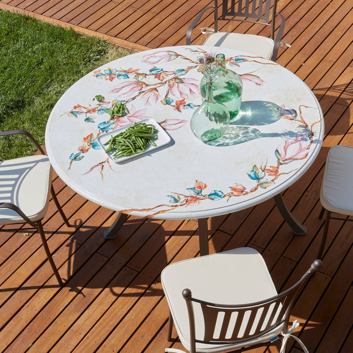 A round outdoor dining table with a white marble-look surface and colorful floral and bird motifs, surrounded by metal patio chairs on a wooden deck. The table holds a green glass bottle and a small tray of greenery.