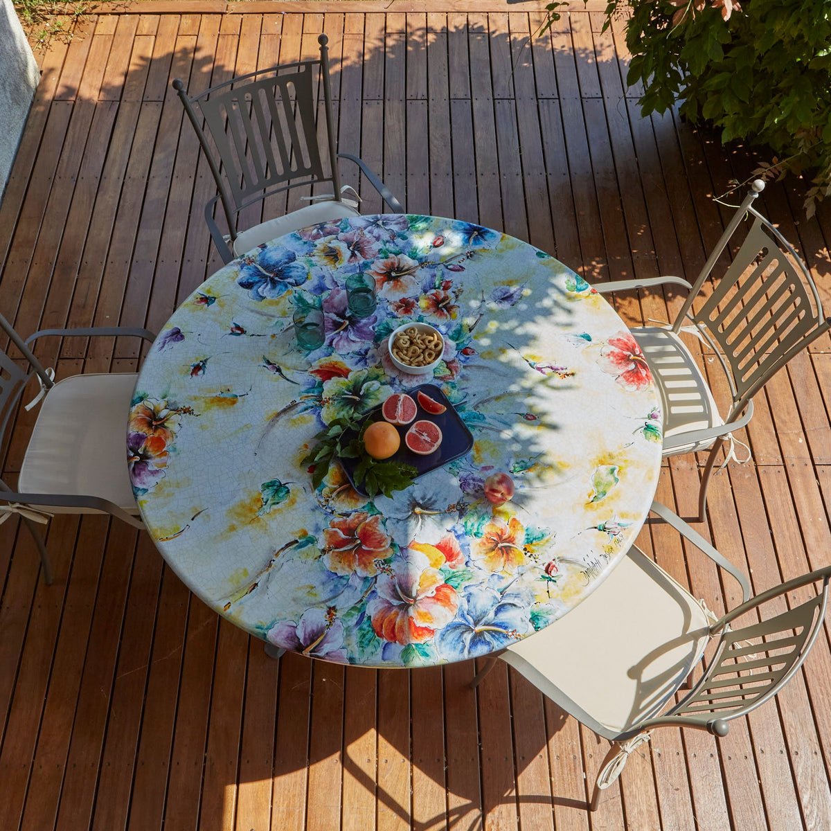 A round outdoor dining table with a colorful floral print, surrounded by four gray metal patio chairs on a wooden deck.