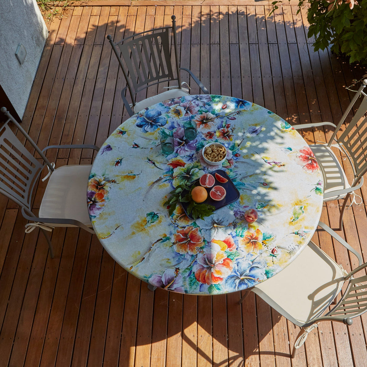 A round outdoor dining table with a colorful floral print, surrounded by four gray metal patio chairs on a wooden deck.