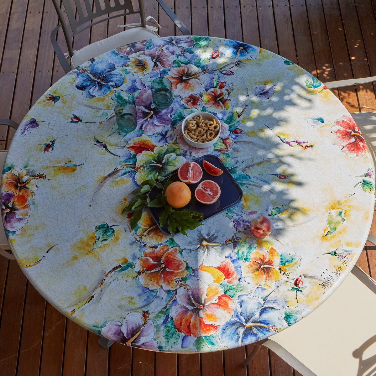 A round outdoor dining table with a colorful tropical floral print, surrounded by metal patio chairs on a wooden deck.