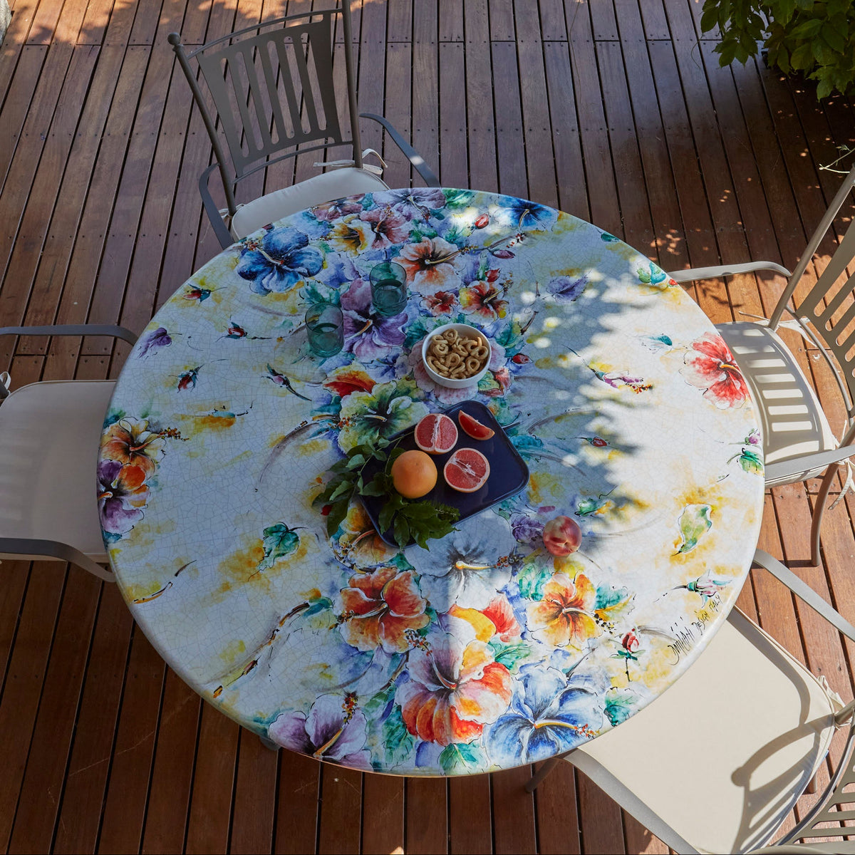 A round outdoor dining table with a colorful floral print, surrounded by metal patio chairs on a wooden deck with green grass and foliage in the background.