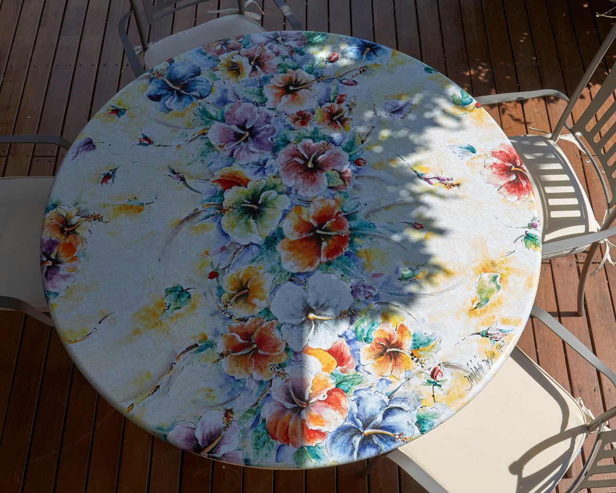 A round outdoor dining table covered with a white tablecloth featuring a colorful floral print, surrounded by metal patio chairs on a wooden deck.