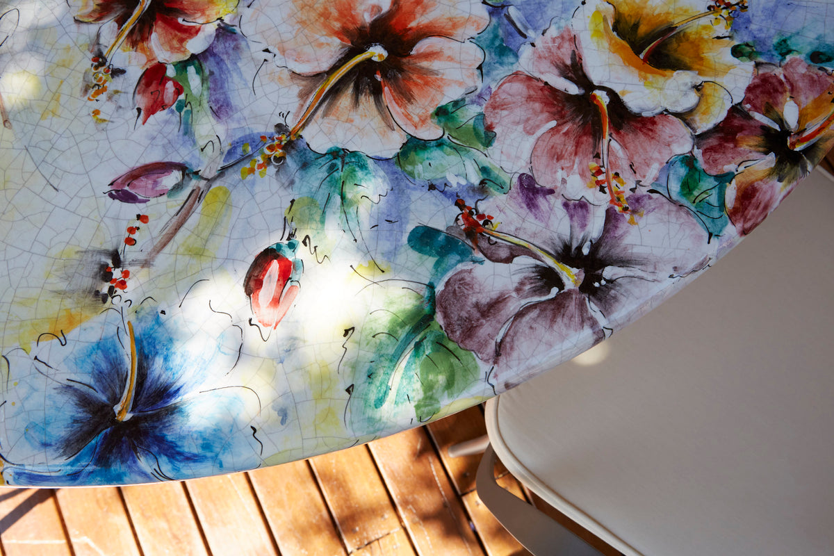 A close-up of a round table or tray featuring a vibrant watercolor floral print with hibiscus flowers in blue, purple, pink, and green on a light background. The table is photographed outdoors on a wooden deck next to a white chair, with dappled sunlight filtering through leaves.