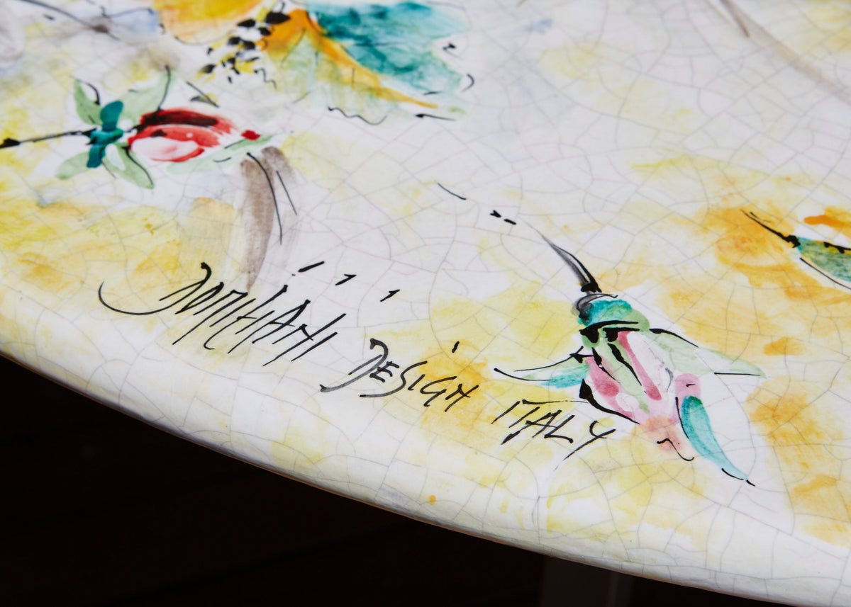 A close-up of a white ceramic table top featuring a hand-painted watercolor design with yellow washes, green and pink insects, and black cursive writing that reads 'MILANO DESIGN ITALY'.