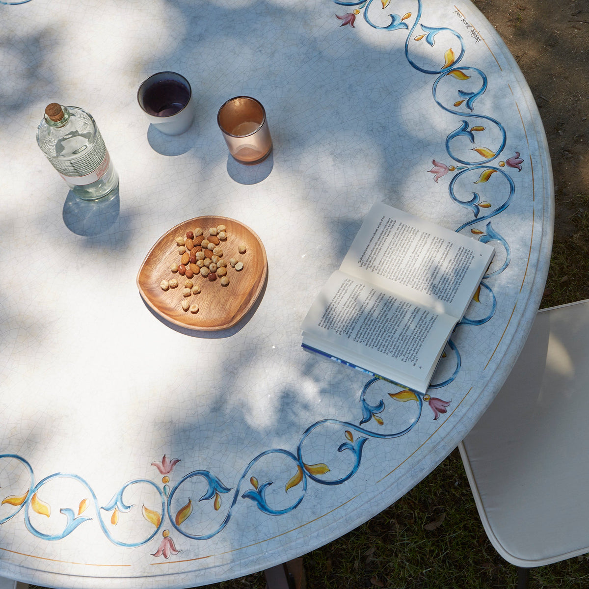 An overhead view of a round outdoor table with a white surface and blue floral scroll border. On the table are a glass bottle, two cups, a small wooden dish with nuts, and an open book, with dappled sunlight casting shadows across the scene.
