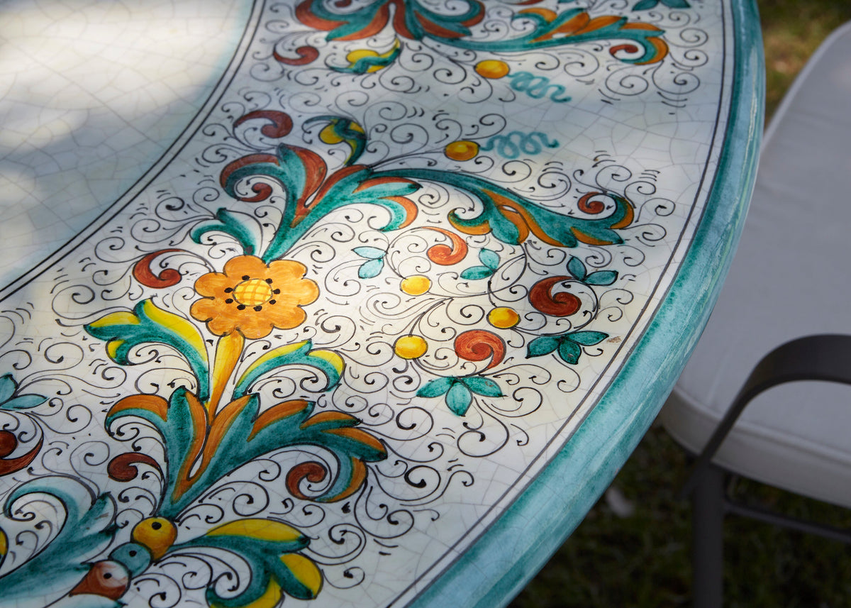 A close-up of a round outdoor table with a white tile surface decorated with colorful hand-painted floral and scroll motifs in teal, orange, yellow, and brown, with a white chair partially visible on the right.