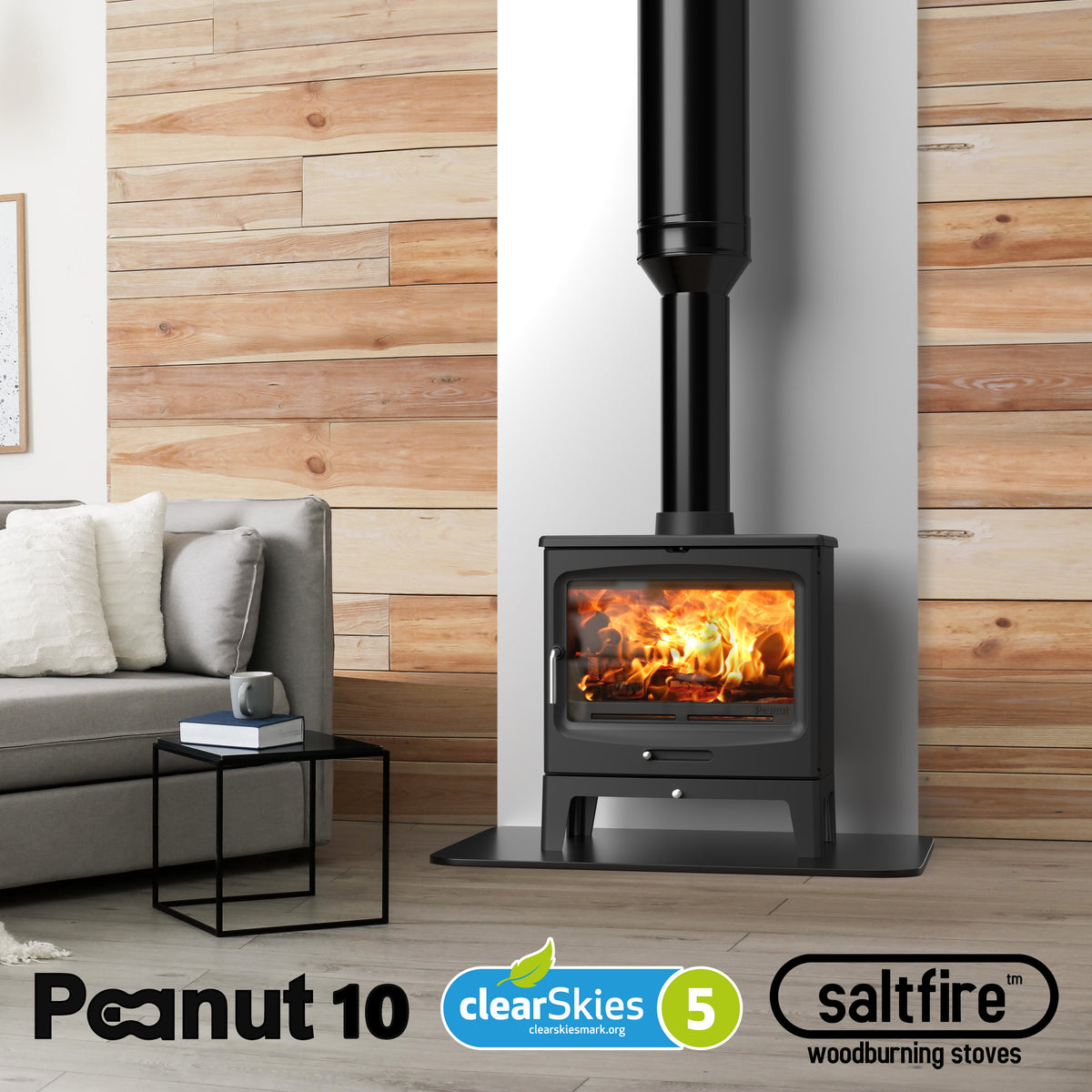 A black Peanut 10 woodburning stove with a glowing fire inside, set in a modern living room next to a gray sofa and a decorated Christmas tree. The image includes 'Peanut 10', 'clearSkies 5', and 'saltfire woodburning stoves' logos at the bottom.