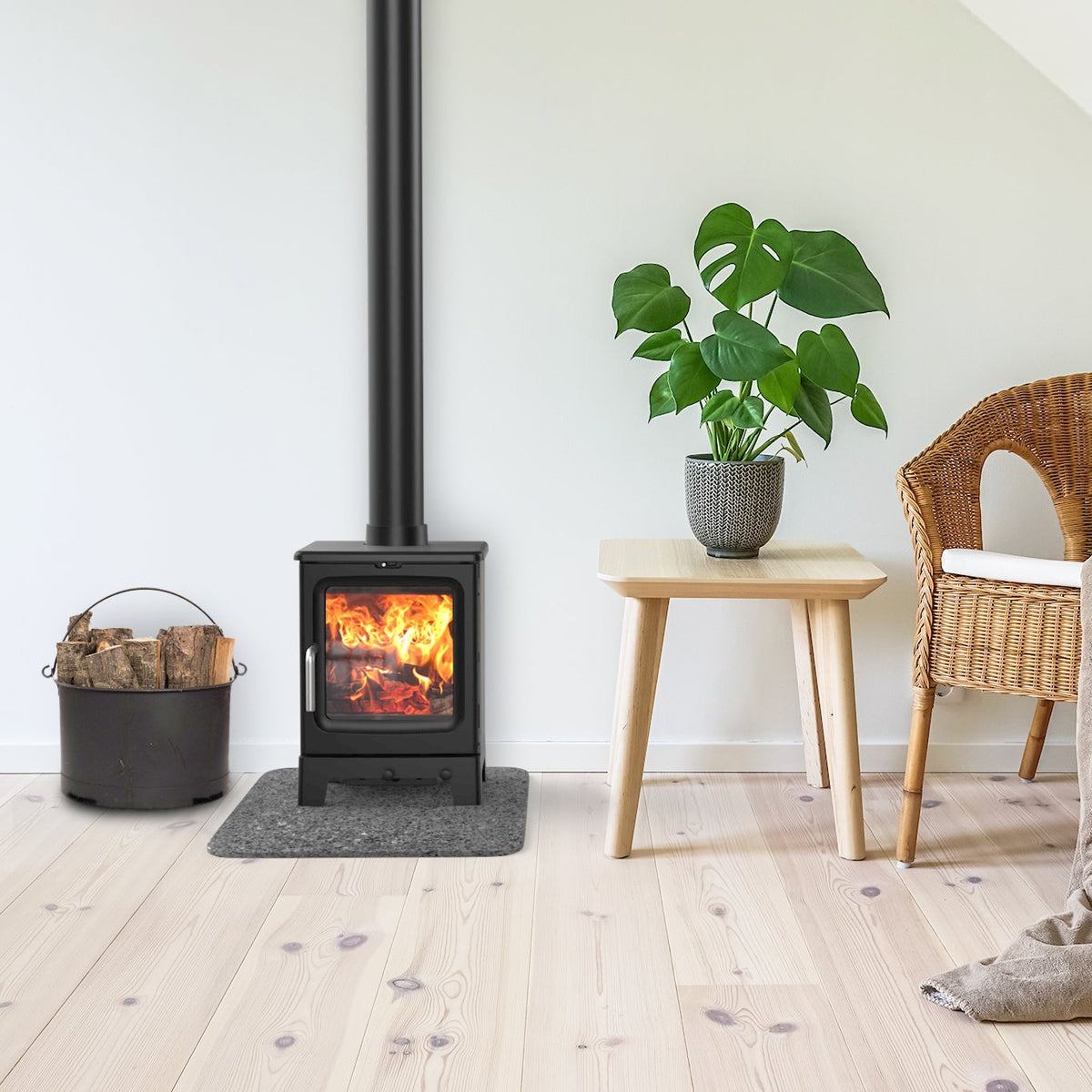 A black wood-burning stove with a visible fire, set in a cozy room with light wood floors, a wicker chair with a beige throw blanket, a small wooden table with a potted green plant, and a bucket of firewood.