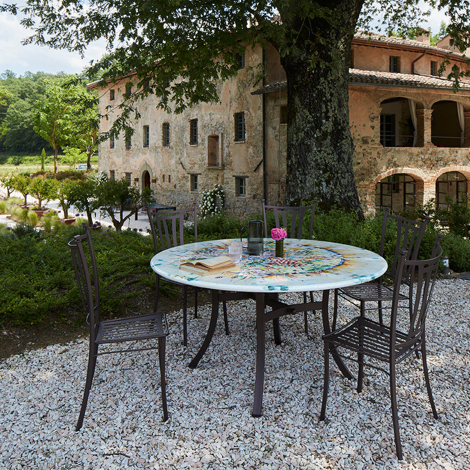 Outdoor patio dining set with a round mosaic table and four dark metal chairs, placed on a gravel area in front of a stone villa with trees in the background.