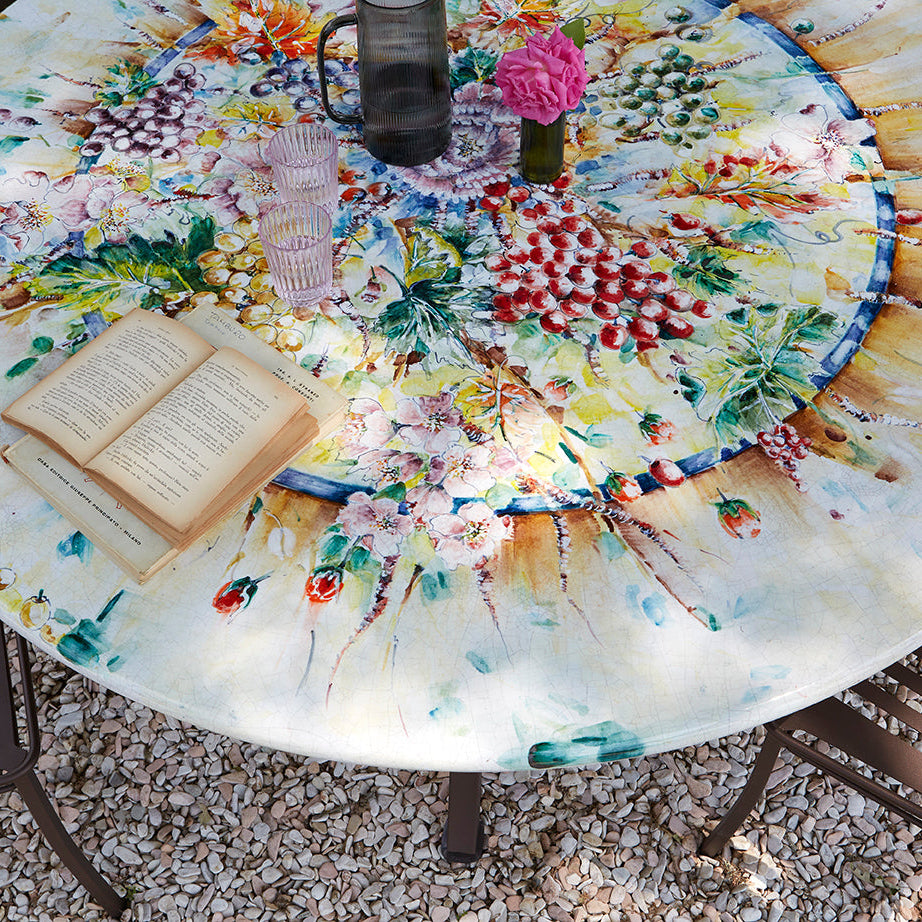 A round outdoor dining table with a highly detailed, colorful floral and botanical print, surrounded by dark metal patio chairs on a gravel surface. The table is styled with an open book, a small vase of pink flowers, a glass, and a dark mug.