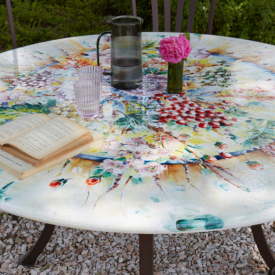 A round outdoor patio table with a colorful floral and abstract painted design on the top, supported by dark metal legs. The table is styled with an open book, a glass, a pink flower in a vase, and a glass pitcher, set on a gravel surface with garden chairs and foliage in the background.