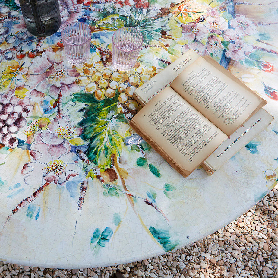 A round outdoor table covered in a colorful floral and fruit print, with an open book and two clear drinking glasses placed on top. The table is set on a gravel surface.