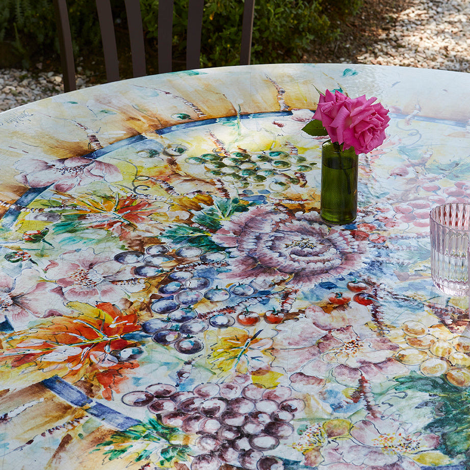 A round outdoor dining table with a vibrant, multicolored floral and botanical print tablecloth, styled with a small green vase holding a pink rose and a glass cup. A dark brown wooden chair is positioned behind the table in a sunny garden setting with green grass and stone steps in the background.