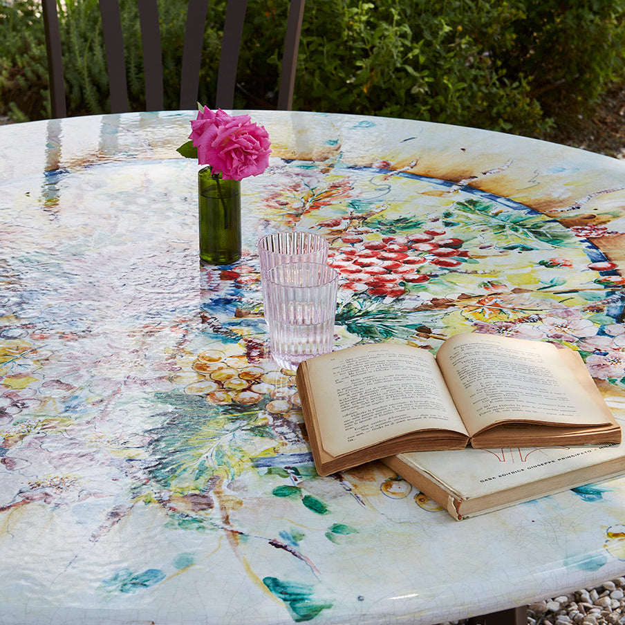 A round outdoor dining table with a colorful, abstract hand-painted mosaic design, styled with an open book, a pink flower in a small green vase, and a clear glass on a patio in a garden setting.