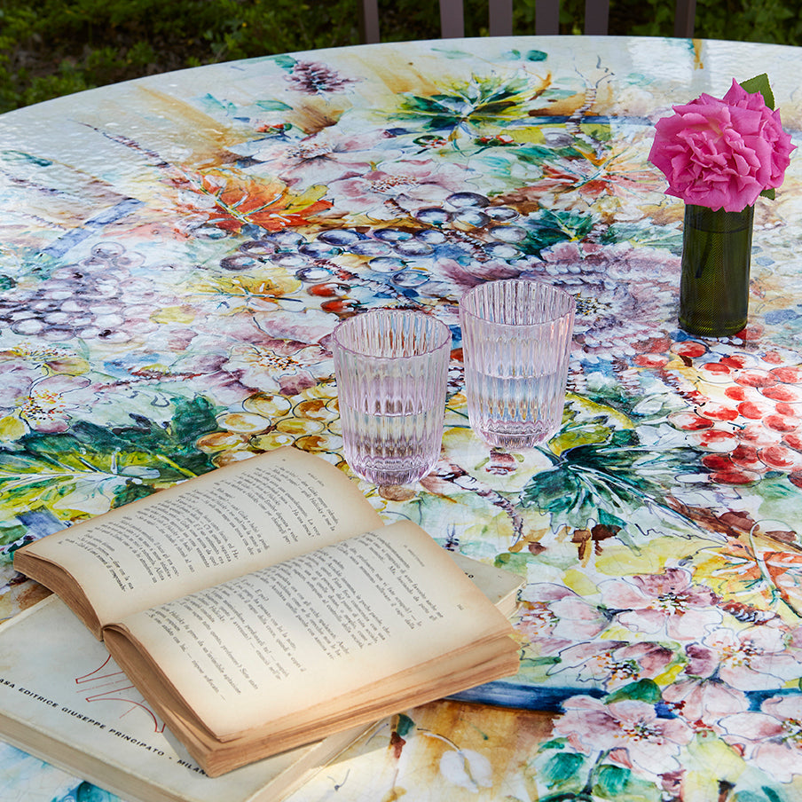 A round outdoor table covered in a colorful floral and botanical print, styled with two pink tinted drinking glasses, an open vintage book, and a small vase of pink flowers. A dark wooden chair is visible in the background against green garden foliage.