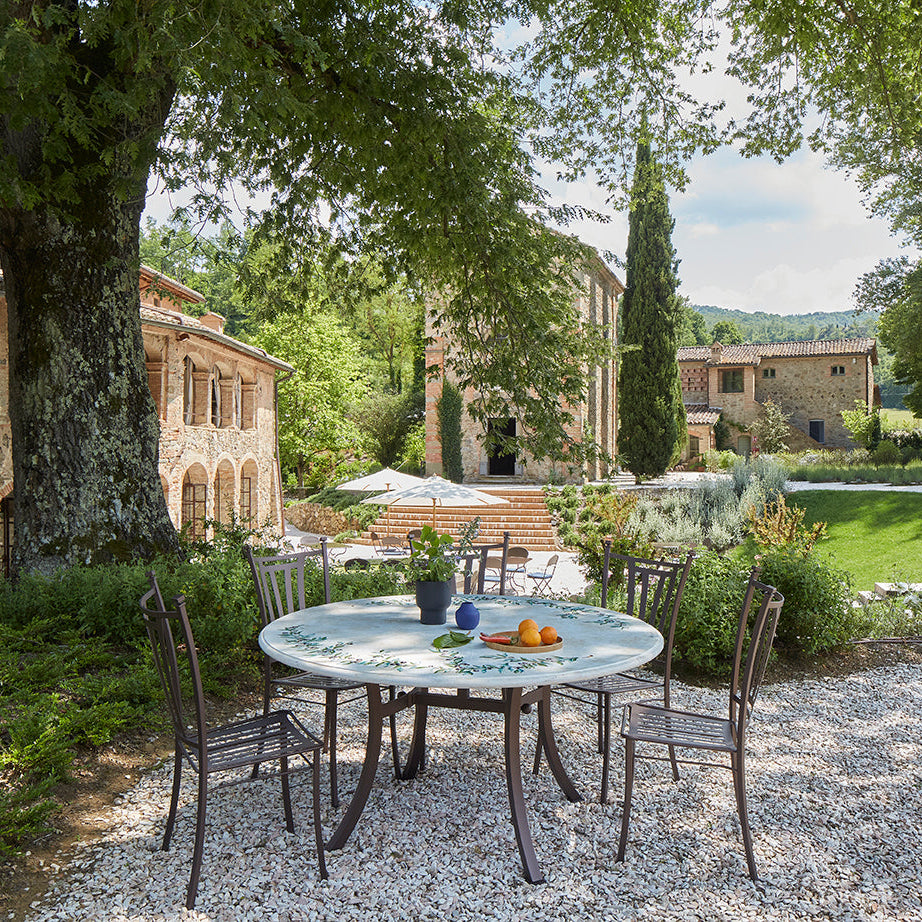 Outdoor dining setup with a round mosaic table and four dark metal chairs on a gravel patio, set in a lush garden courtyard with stone buildings, trees, and a lawn in the background.