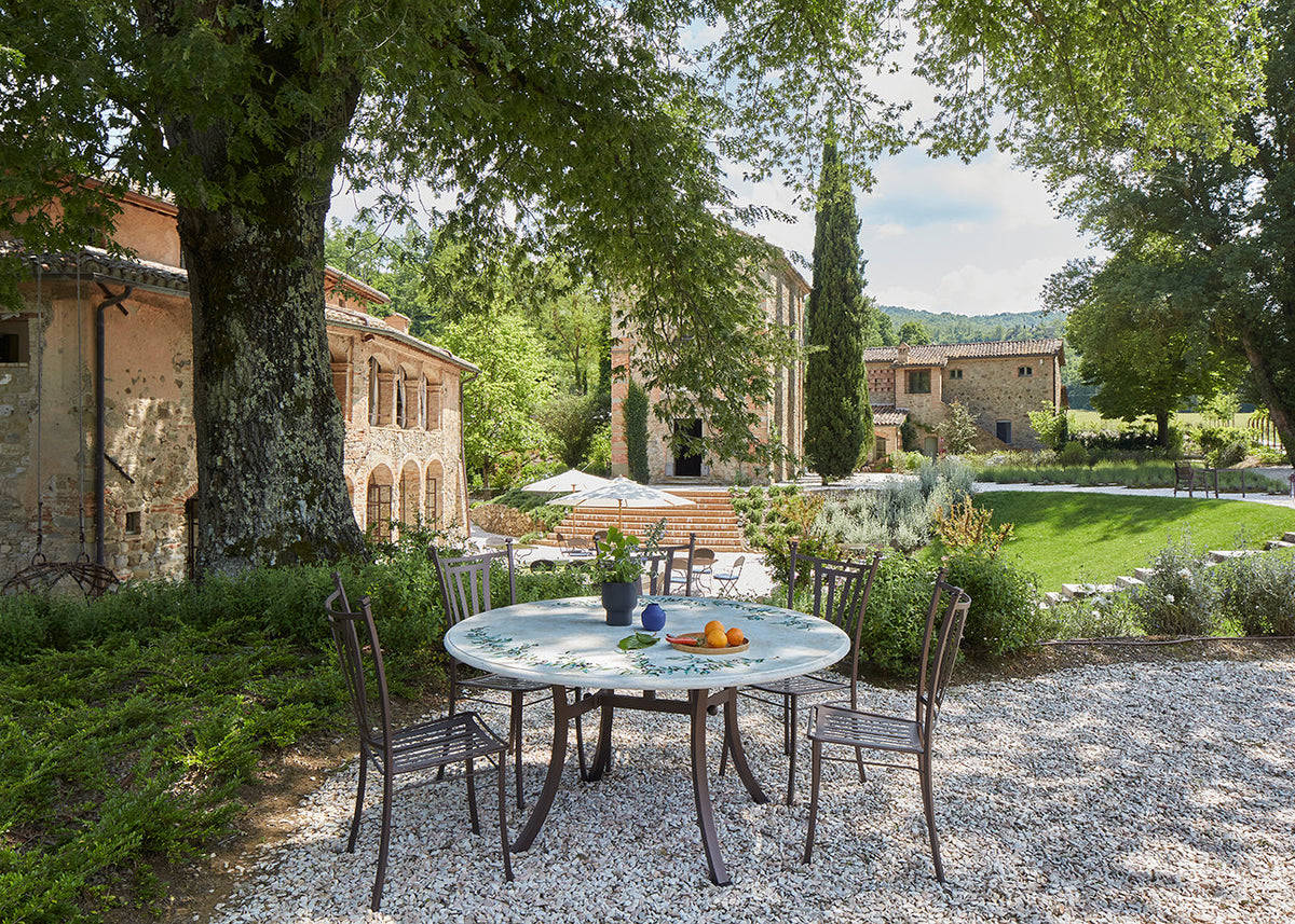 Outdoor dining setup with a round mosaic table and four dark metal chairs on a gravel patio, set in a lush garden courtyard with stone buildings, trees, and a lawn in the background.