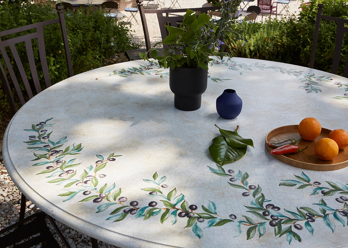 A round outdoor dining table with a white stone or concrete surface decorated with painted green leaf and berry motifs around the edge. The table is styled with a black vase of greenery, a small blue vase, a wooden tray with oranges, and leaves.
