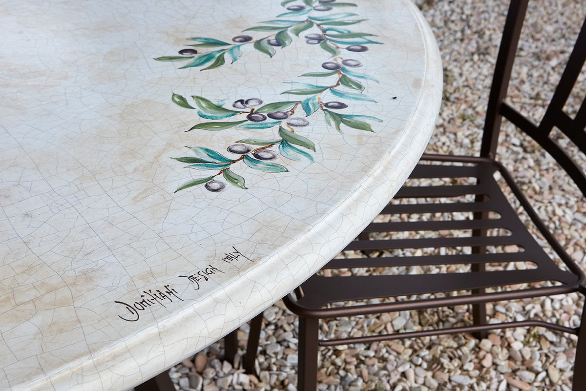 A close-up of a round outdoor table with a distressed cream-colored ceramic tile surface, decorated with a painted green and purple olive branch motif. A dark metal patio chair is partially visible next to the table on a gravel surface.